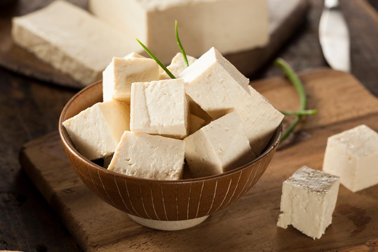 Sliced tofu blocks are stacked in a wooden bowl with other slices of tofu laying beside it on a wooden cutting board.