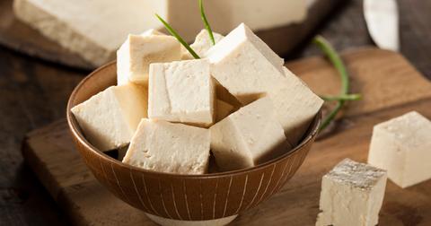Sliced tofu blocks are stacked in a wooden bowl with other slices of tofu laying beside it on a wooden cutting board.
