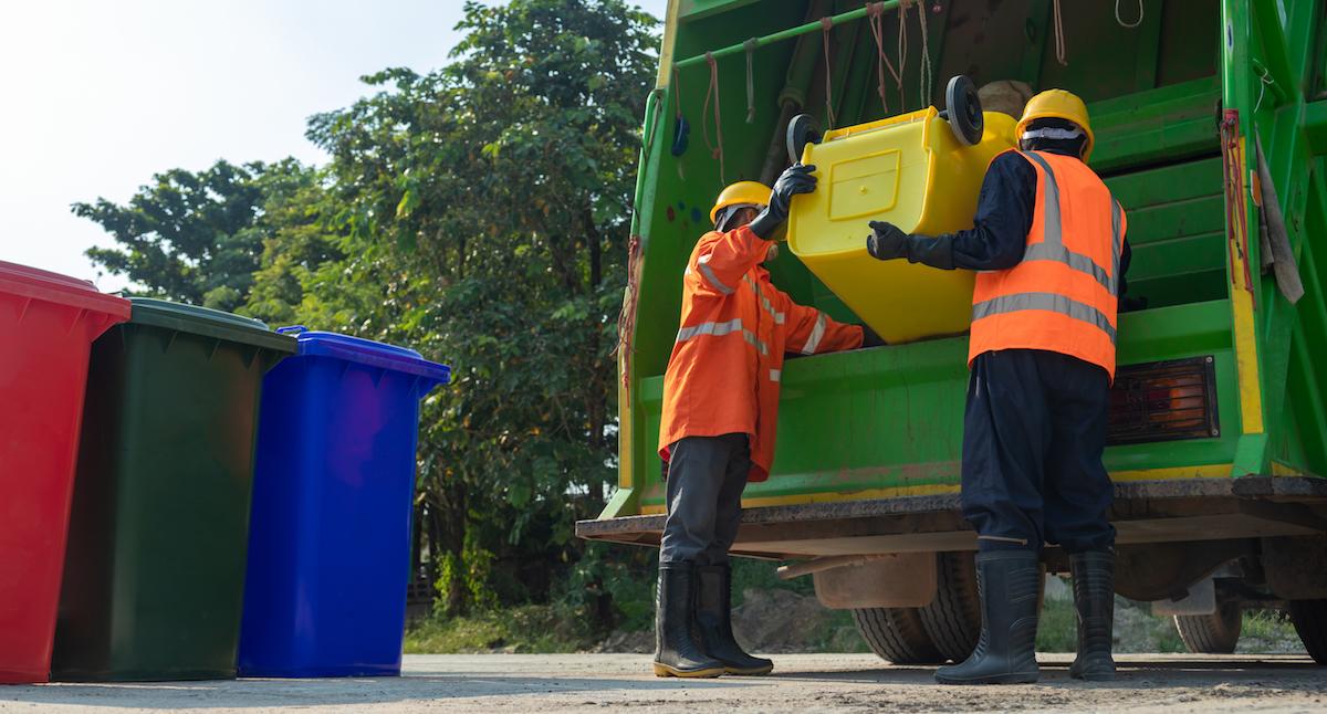 Two sanitation workers pour a bin into a garbage truck