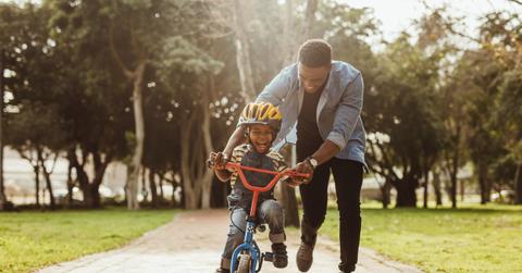 A smiling father steadies his smiling young son on a bicycle in the park.
