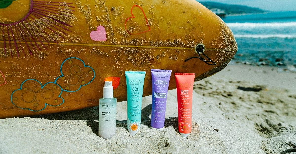 Photo of four Pacifica brand skincare products sitting in the sand in front of a propped up surfboard