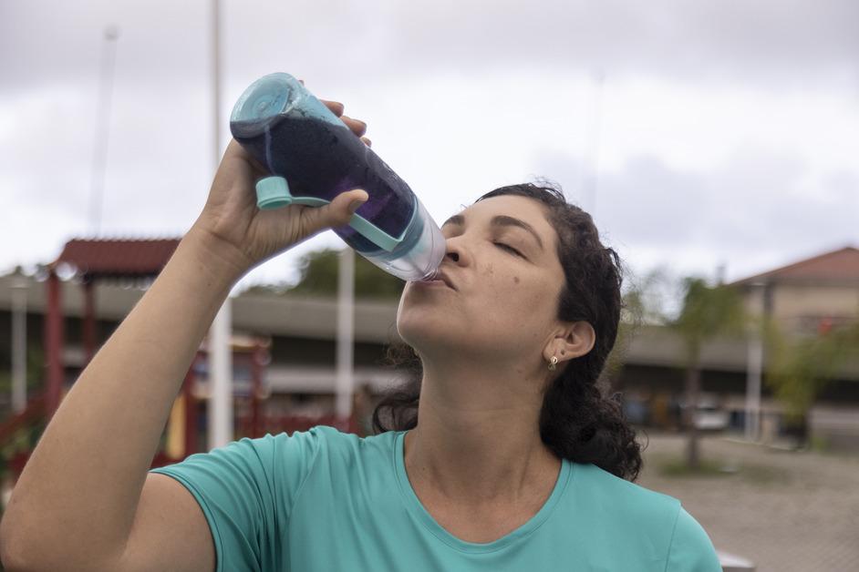 Woman with dark curly hair wearing a teal shirt stands outside while drinking from a water bottle filled with purple liquid. 