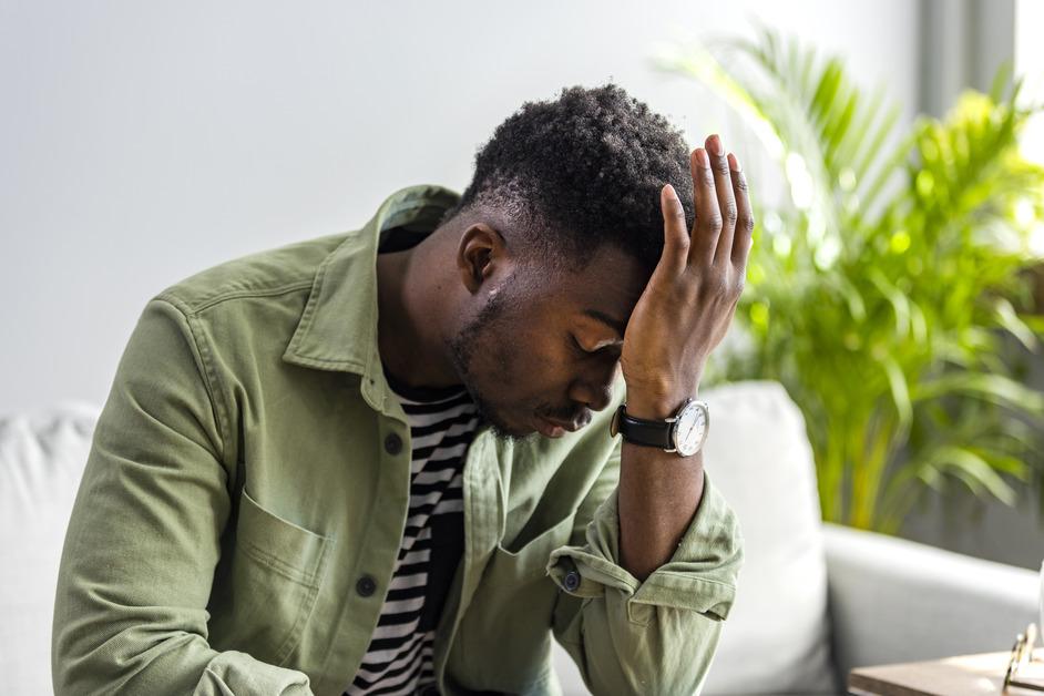 A man in a green jacket and a white and black striped shirt sitting on the couch with his head in his hand looking distressed. 