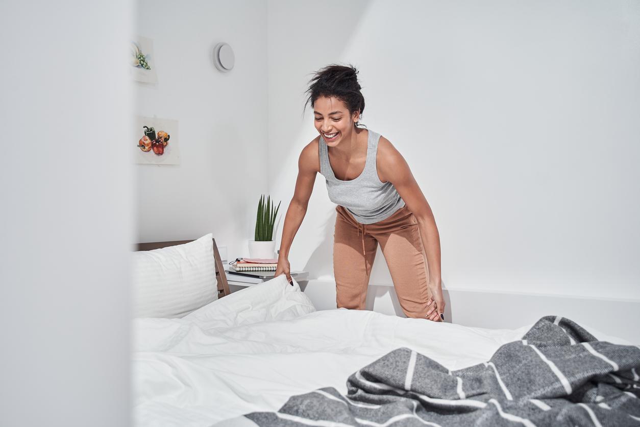 A smiling woman makes the bed by placing a white sheet and gray throw blanket over her bamboo mattress.