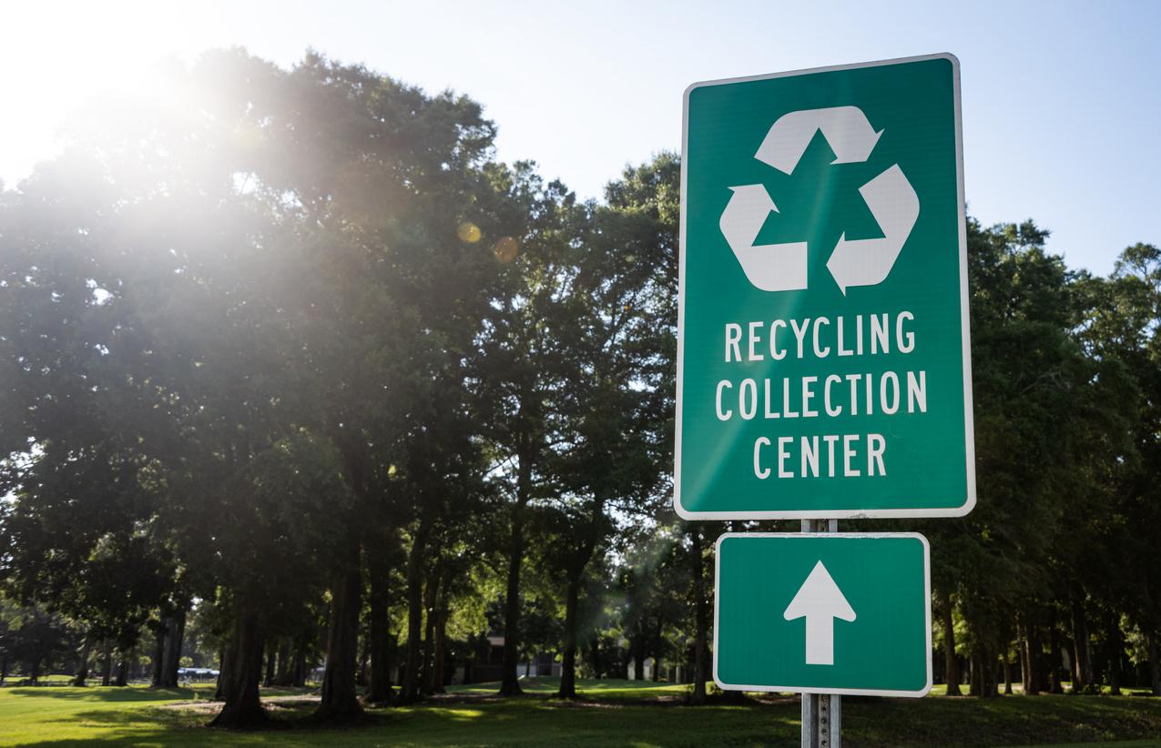 A green sign is pictured in front of large trees, which reads: "Recycling Collection Center" with a recycling symbol and a sign below it with a forward-facing arrow.