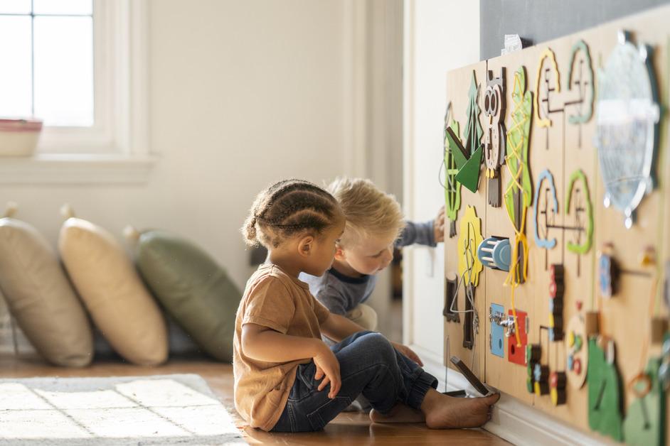 Two children play at a wooden stimulation wall in a room with pillows in the background. 