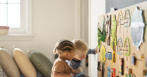 Two children play at a wooden stimulation wall in a room with pillows in the background.