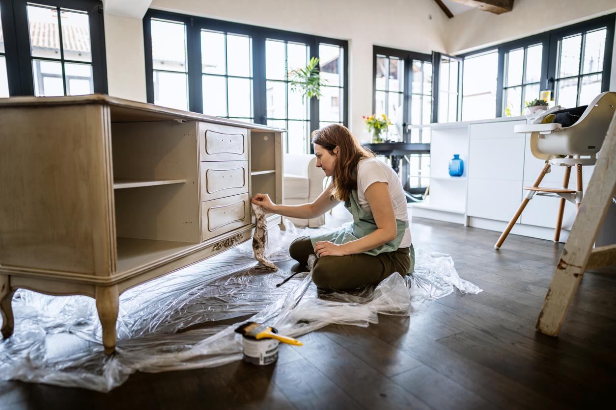 A woman wearing an apron restores a piece of wooden furniture atop a plastic floor covering inside a well-lit home.