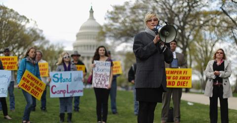 Woman with a megaphone stands in front of protestors in front of the White House with signs that say "Stand with Camp Lejeune."