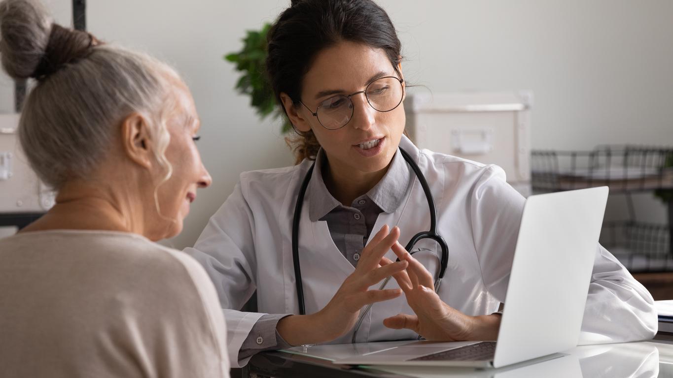 A female doctor in a white coat with a stethoscope explains information to an older female patient while reading information from a white laptop