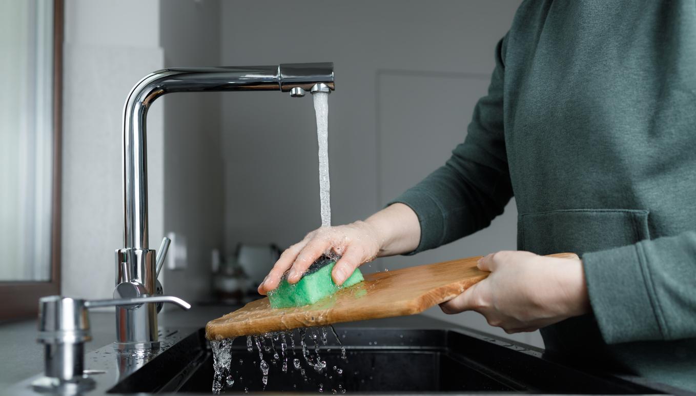 An individual cleans a wooden cutting board with a sponge under running water in a kitchen sink.