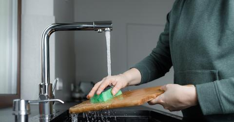 An individual cleans a wooden cutting board with a sponge under running water in a kitchen sink.