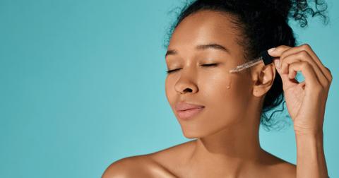 Photo of woman dropping a skincare serum on her cheek in front of a blue background