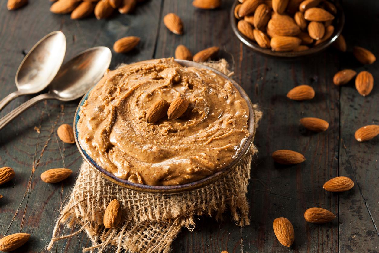 Raw organic almond butter is pictured in a bowl atop a burlap cloth on a wooden table with almonds scattered about.