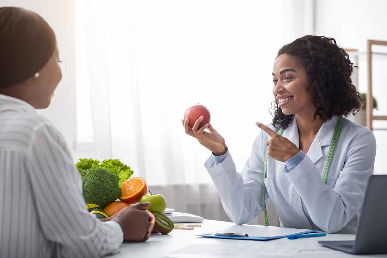 A dietitian points to an apple while smiling at her client sitting across the desk. Fruits and vegetables are stacked beside them.