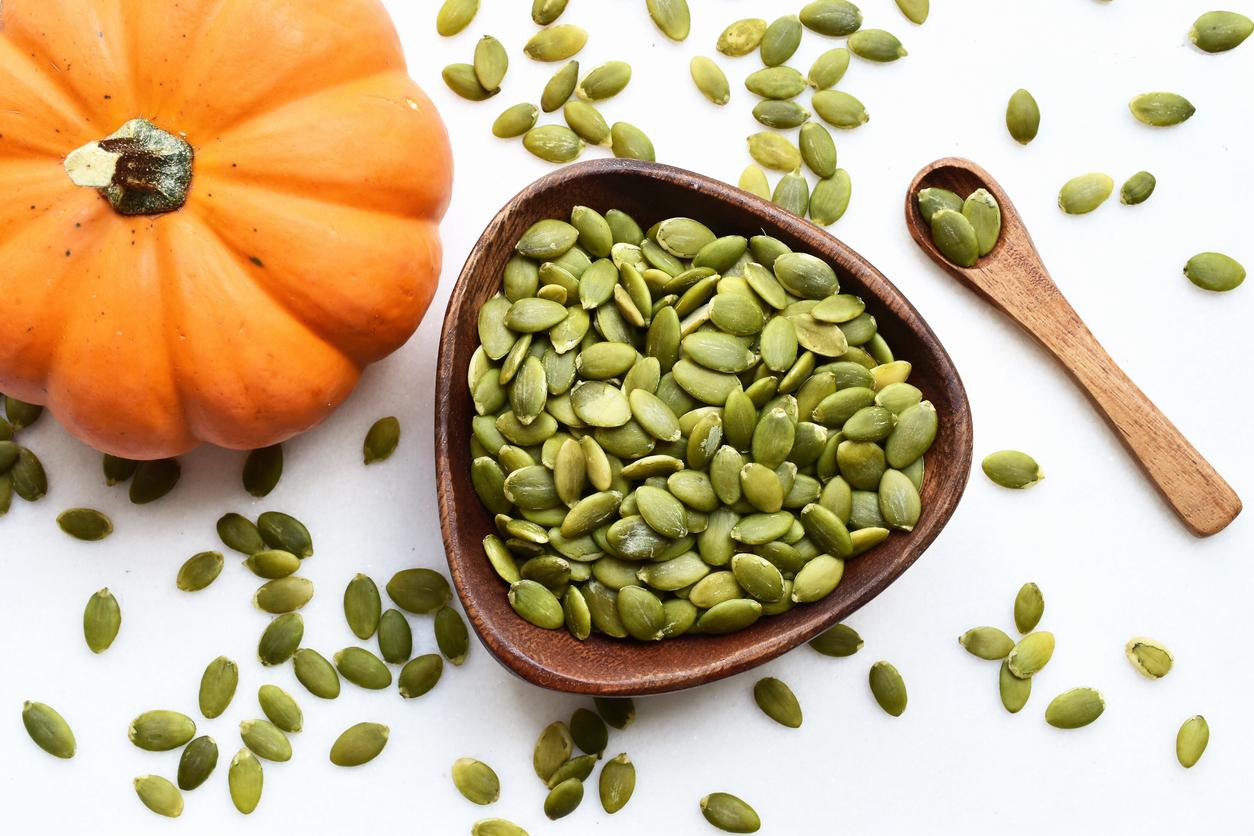 A bowl of pumpkin seeds is pictured beside a small pumpkin and a wooden spoon.