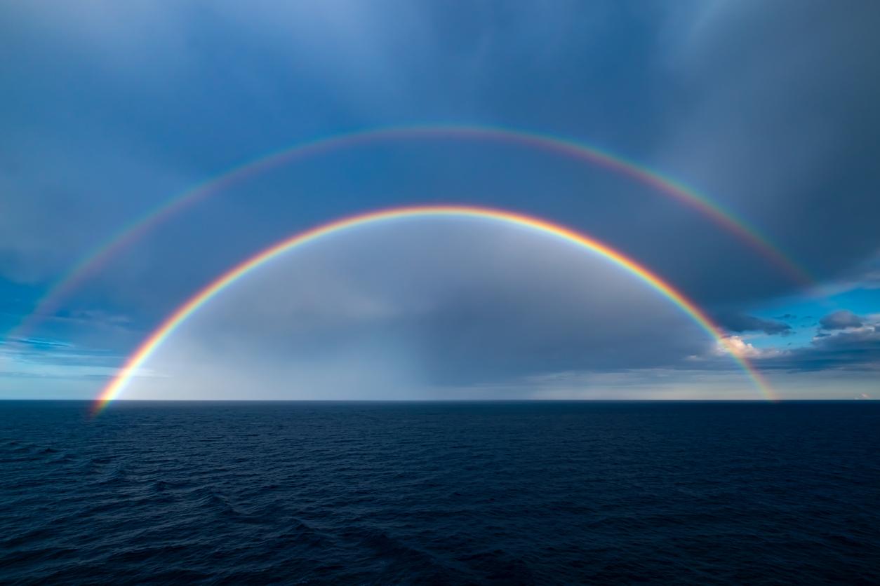 A double rainbow appears over an ocean with cloudy skies behind the double rainbow.