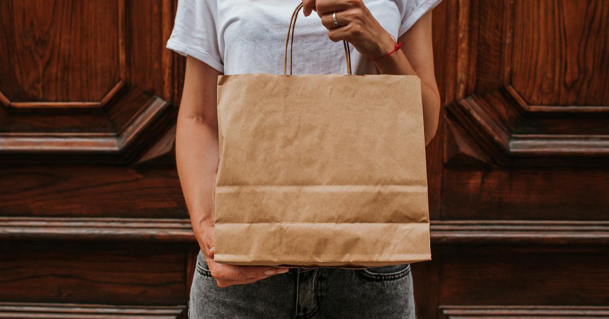 Woman stands in front of a woman door with a paper shopping bag.