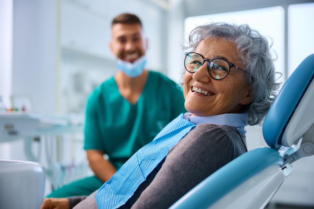 A smiling woman with a blue bib lays in a dentist chair while a dental hygienist smiles at her in the blurry background.