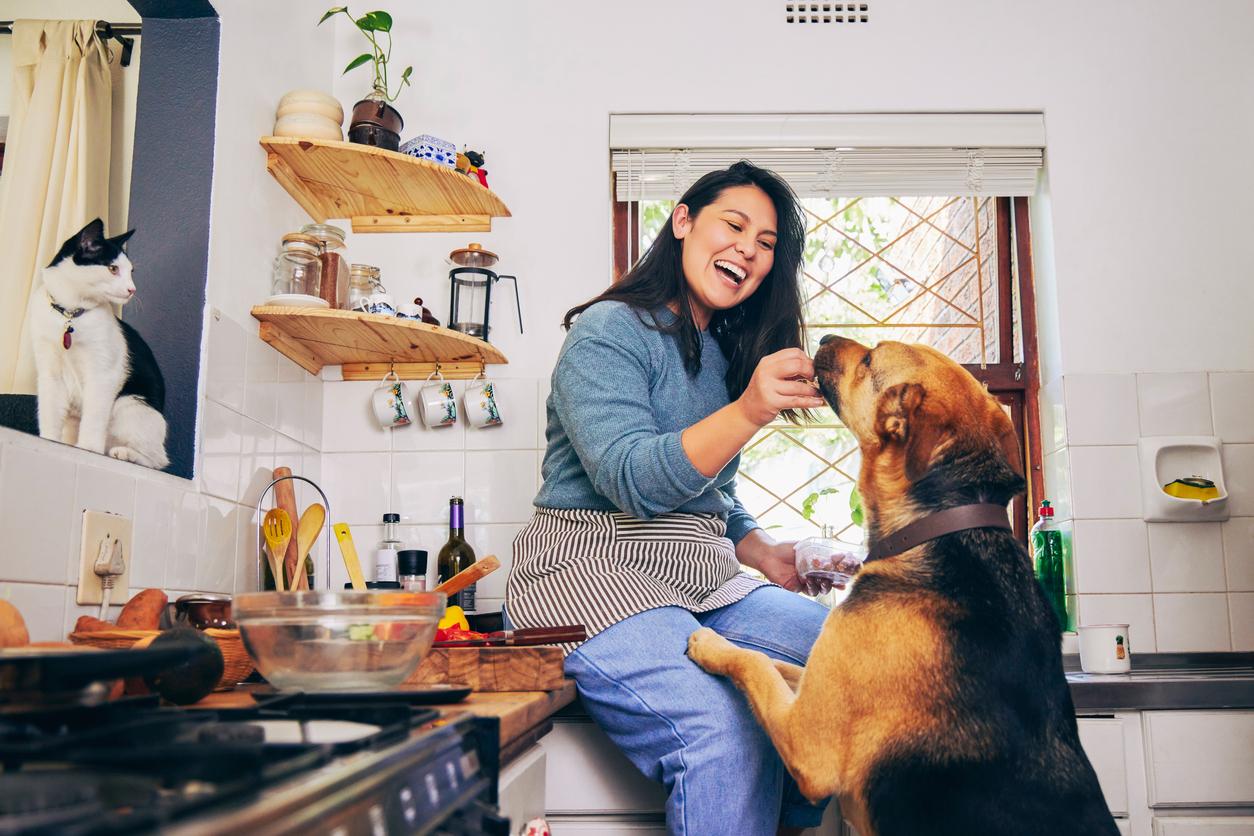 A smiling woman feeds her dog in a kitchen.