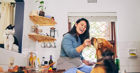 A smiling woman feeds her dog in a kitchen.