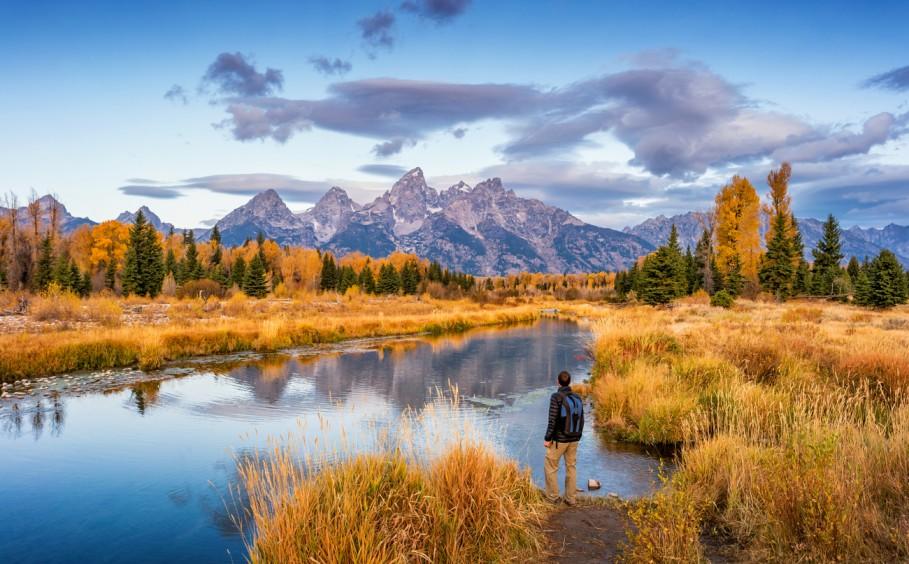 A hiker in Grand Teton National Park admires the autumn colors of the trees beside a river and mountains.