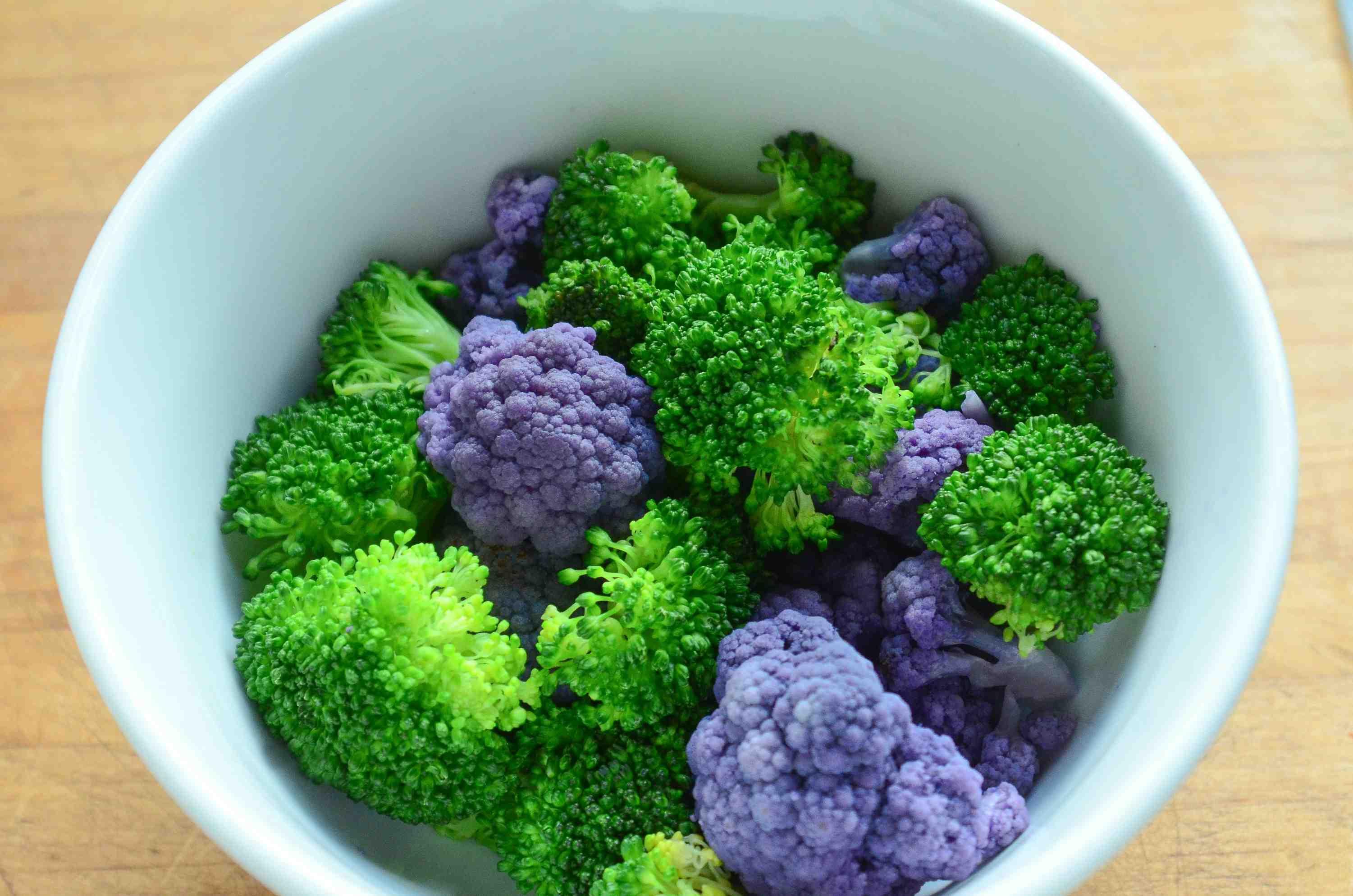 Steamed broccoli and purple cauliflower are pictured in a white bowl.