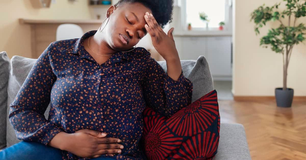 Someone sitting on a sofa with one hand on their abdomen, the other on the forehead with eyes closed.