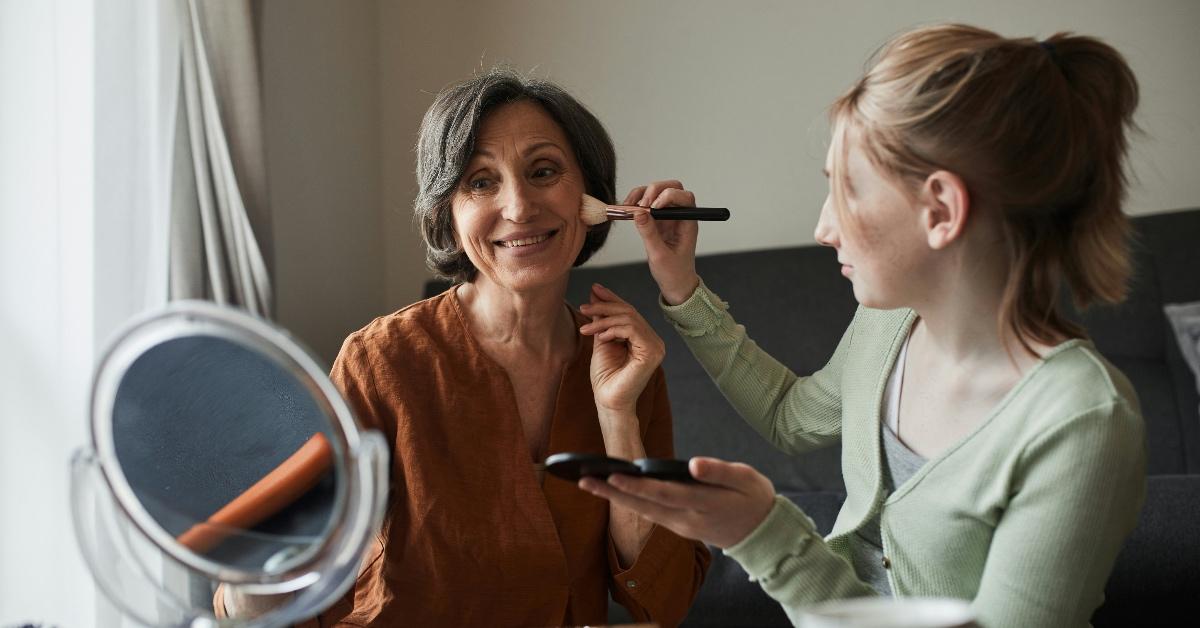 A younger woman in a green sweater helps apply makeup to the face of an older woman in an orange wrap top.