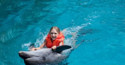 A smiling young girl in an orange life preserver rides on the back of a dolphin.
