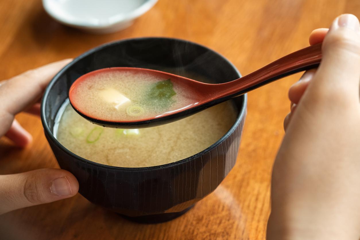 A person holds a black bowl of miso soup in one hand and a red spoon in another.