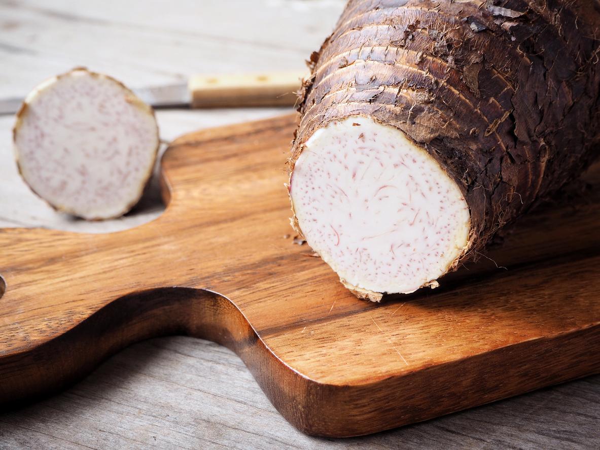 Two taro roots are on a table, one of which is atop a wooden block with a knife in the background.