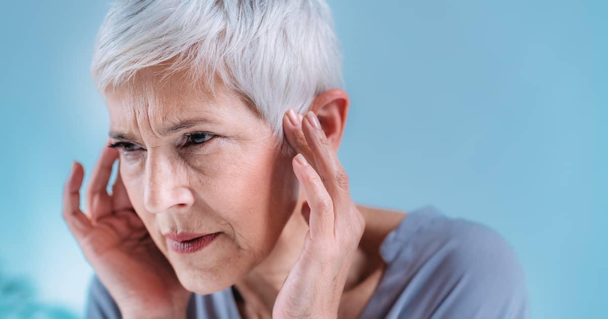 Older person with short grey hair holding her hands up to the side of her head frowning.