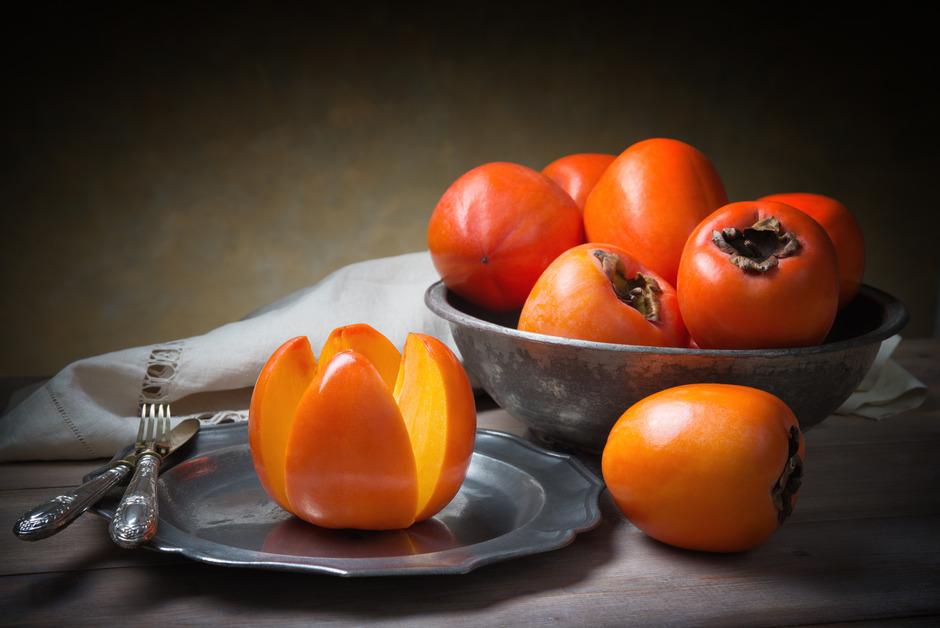 A set of old metal utensils next to a plate and bowl full of persimmons with a white napkin in the background. 