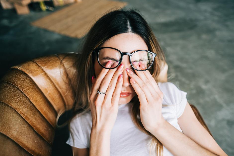 A woman sits in a chair and rubs her eyes under her glasses. 