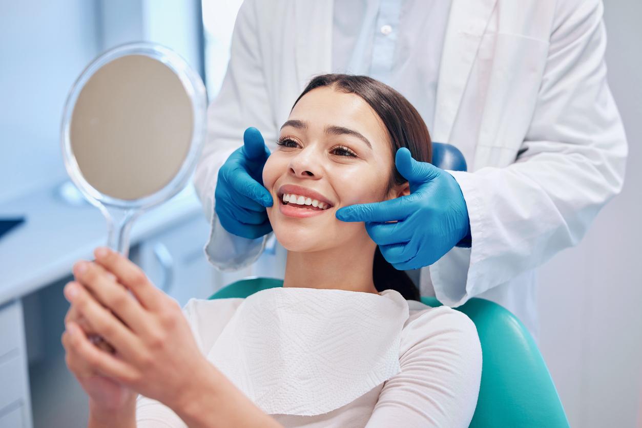 A smiling woman with a white bib lays in a dentist chair while the dentist touches the sides of her mouth with gloved hands.