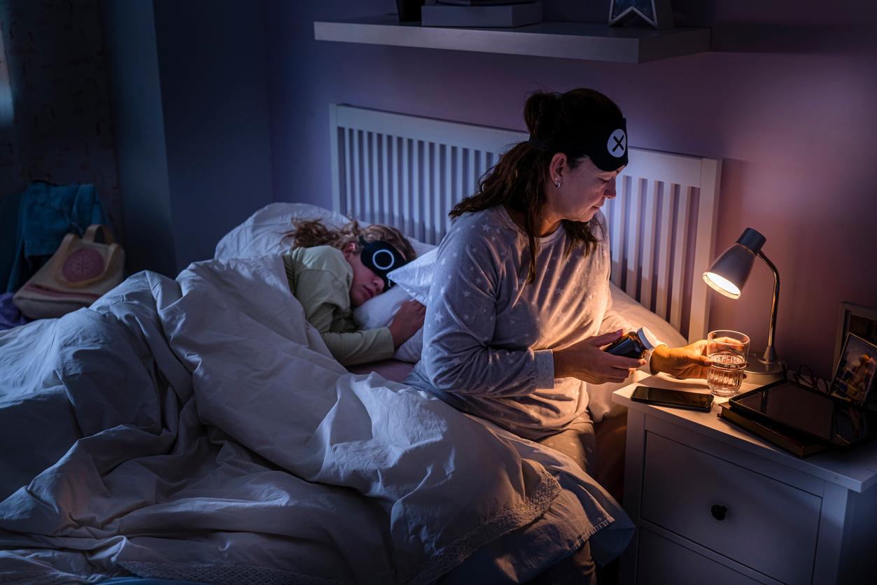 A mother in bed with a sleeping mask on her forehead holds a pill bottle in one hand and a glass of water in another hand.