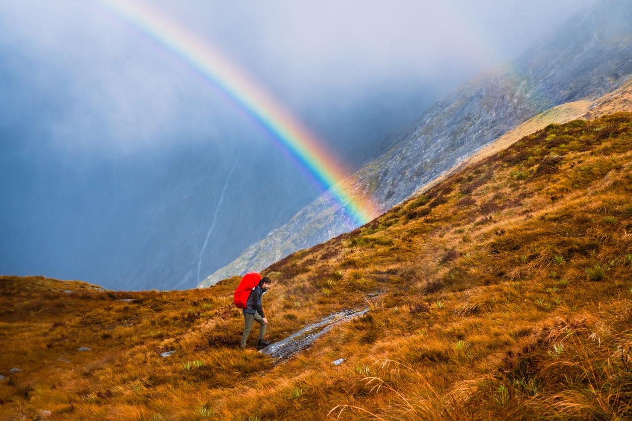 A hiker in red traverses a mountain with a rainbown in the background.