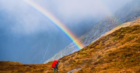 A hiker in red traverses a mountain with a rainbown in the background.