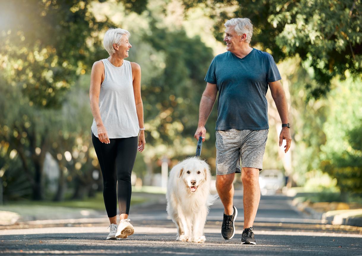 Two older adults in exercise clothes walk their smiling dog while exercising