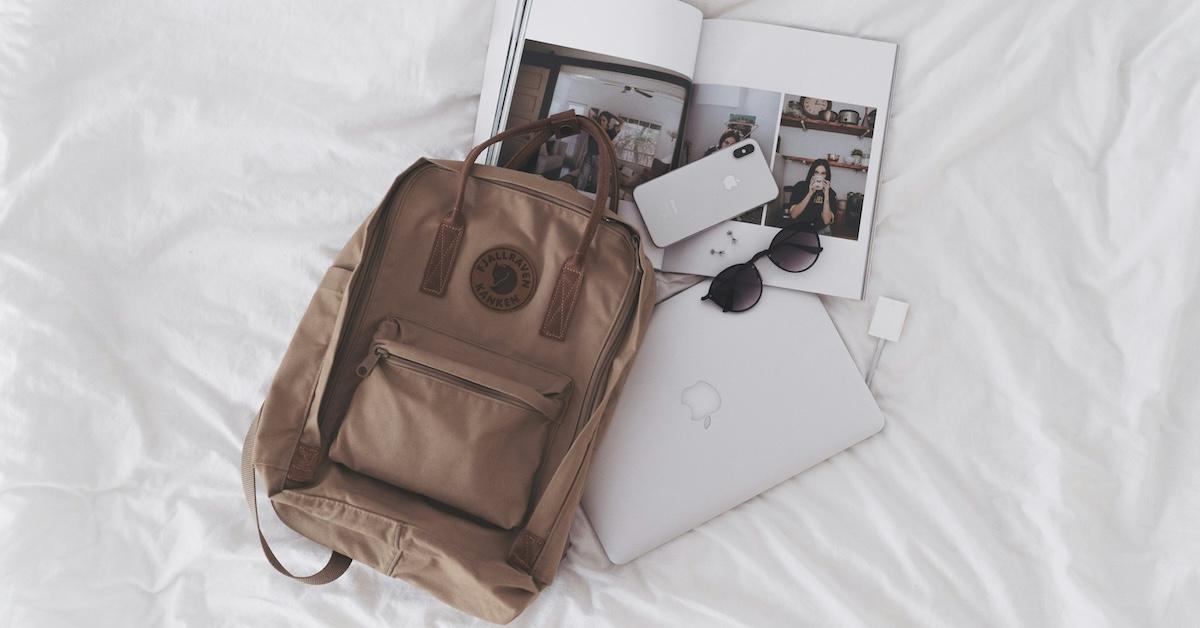 Beige Fjällräven backpack, laptop, phone, magazine, and sunglasses on a bed