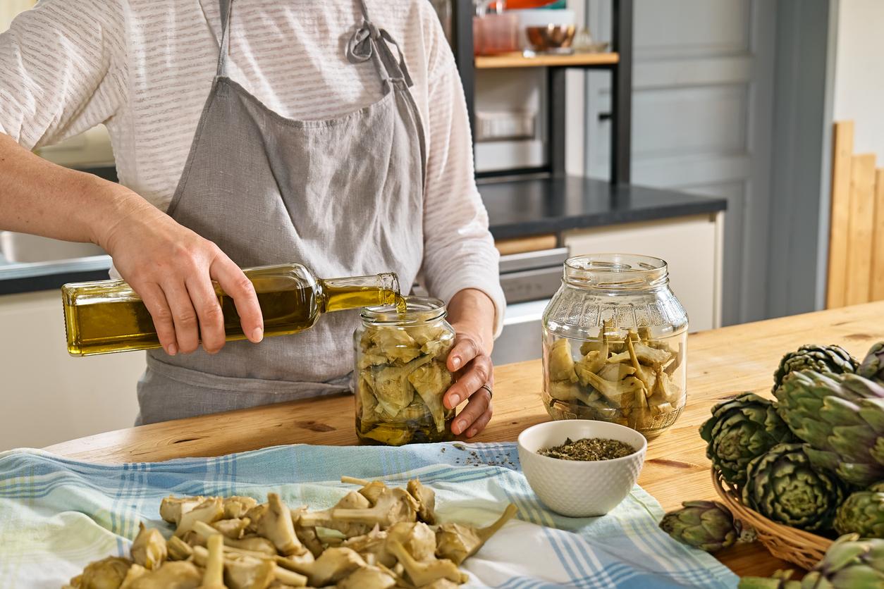 A cook wearing a gray apron pours olive oil into a glass jar of artichoke hearts beside another jar and stacked artichokes on a wooden table.