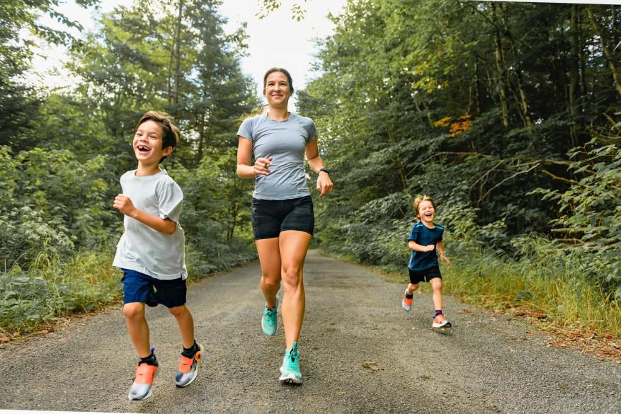 A mother and her two children smile as they run on a dirt trail surrounded by trees.