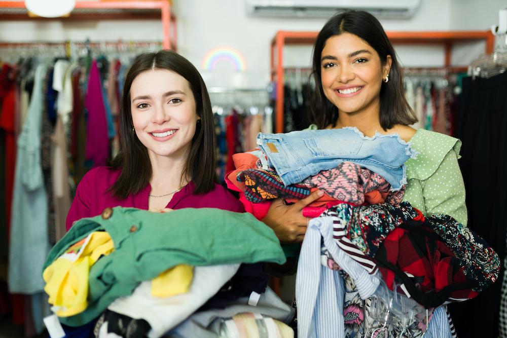 Friends at a thrift store smiling holding clothes.
