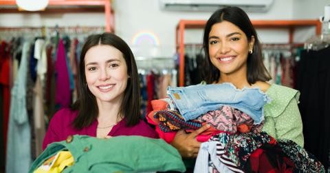 Friends at a thrift store smiling holding clothes.