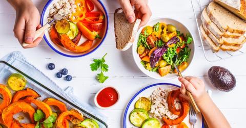 A variety of plant-based meals on a white wooden table, taken from above, with two people's hands reaching in with forks.