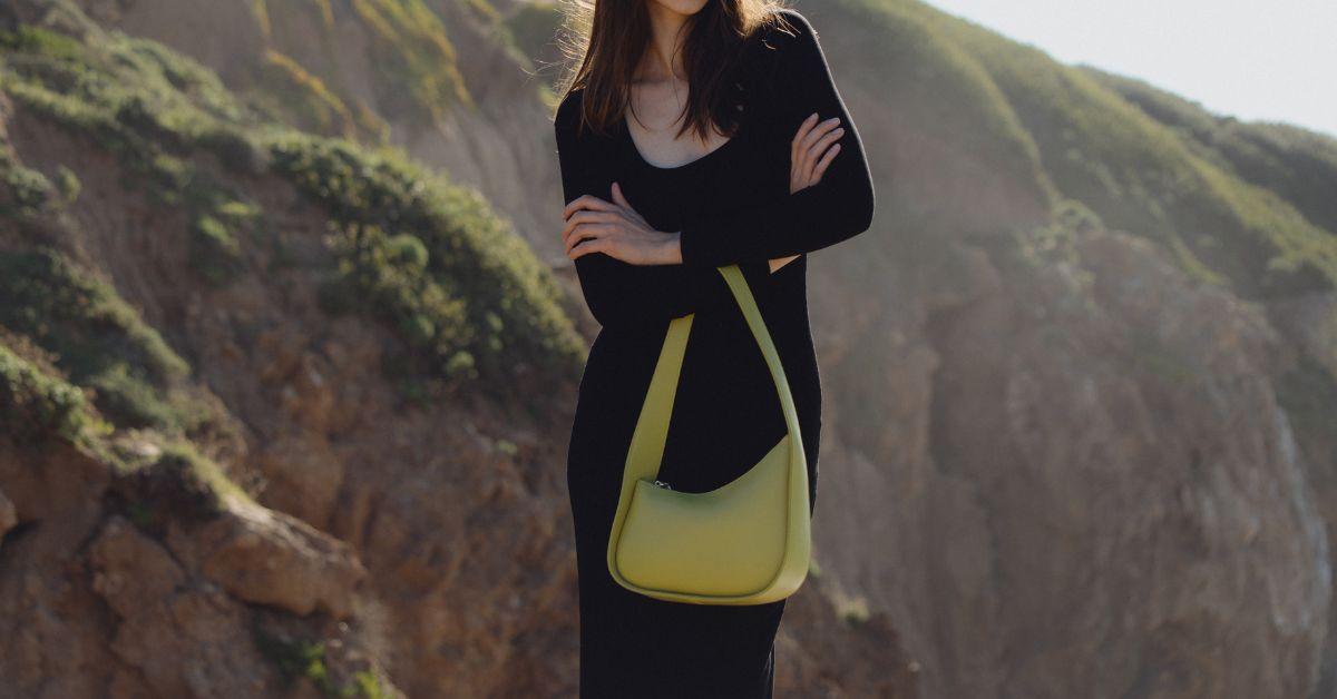 In front of a mountain, a model in a black outfit holds light green handbag by Melie Bianco.