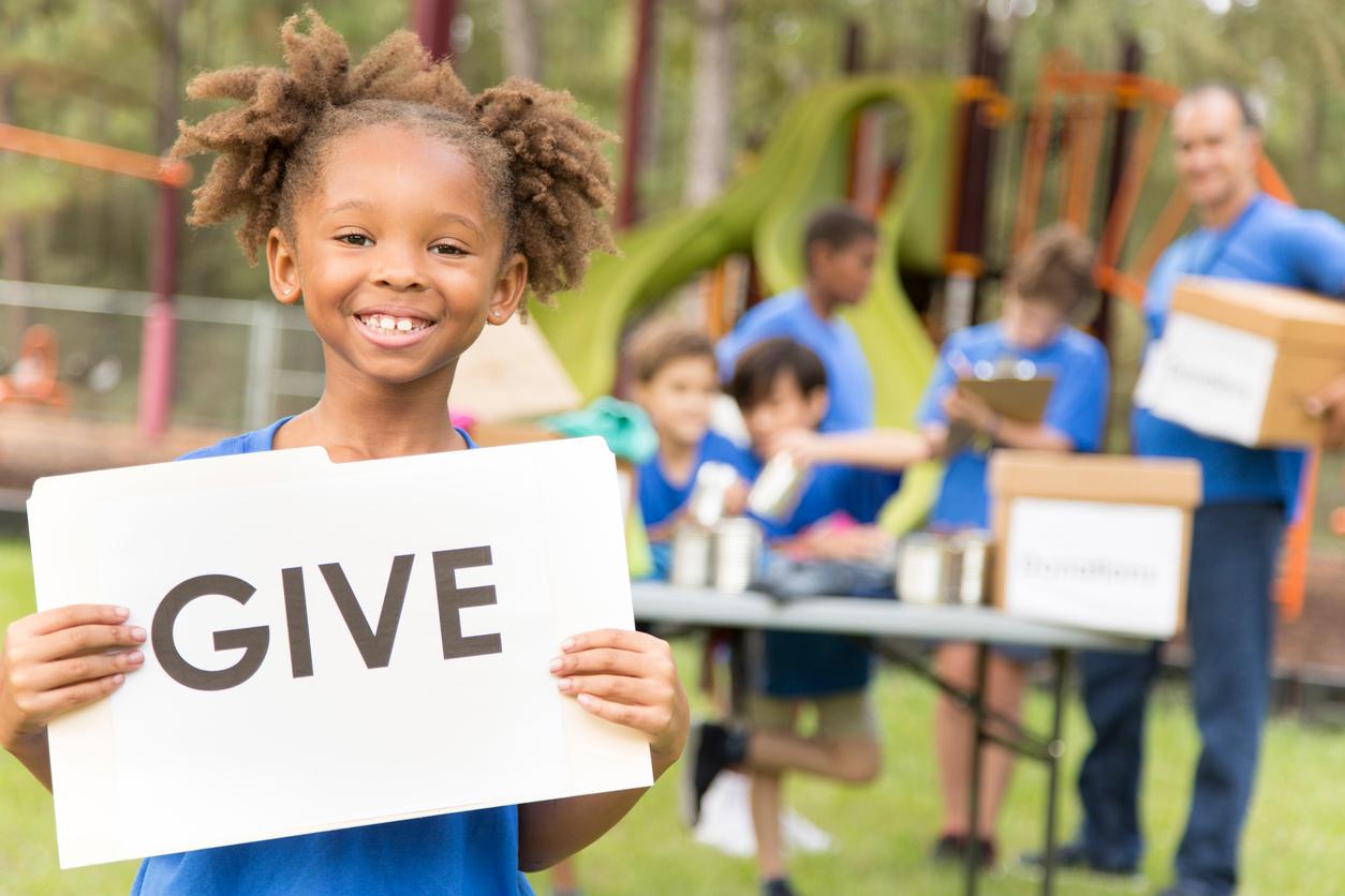 A young child smiles as she holds up a sign that reads "GIVE" with volunteers in the background working a donation drive.