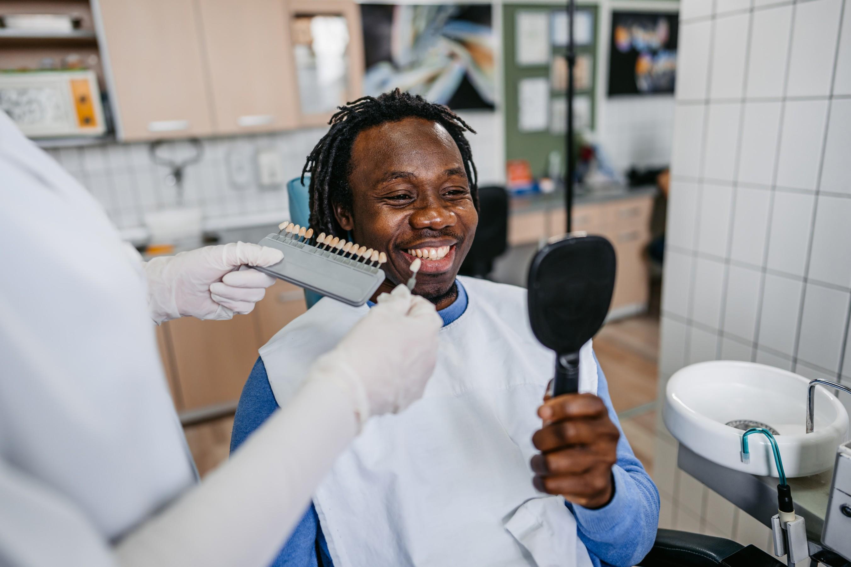 A dentist wearing white gloves uses a palette tooth color sample to match the color of a patient's teeth.