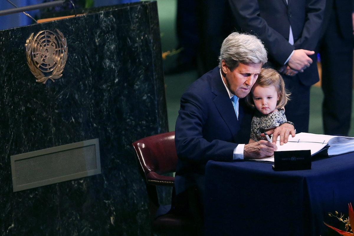 A gentleman in a suit at the UN holds his grand daughter as he signs the Paris Agreement sitting down at a blue table.